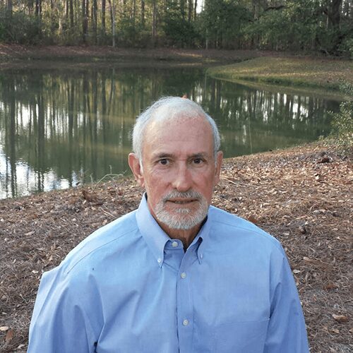 Elderly man wearing a blue shirt standing outdoors near a pond surrounded by trees.