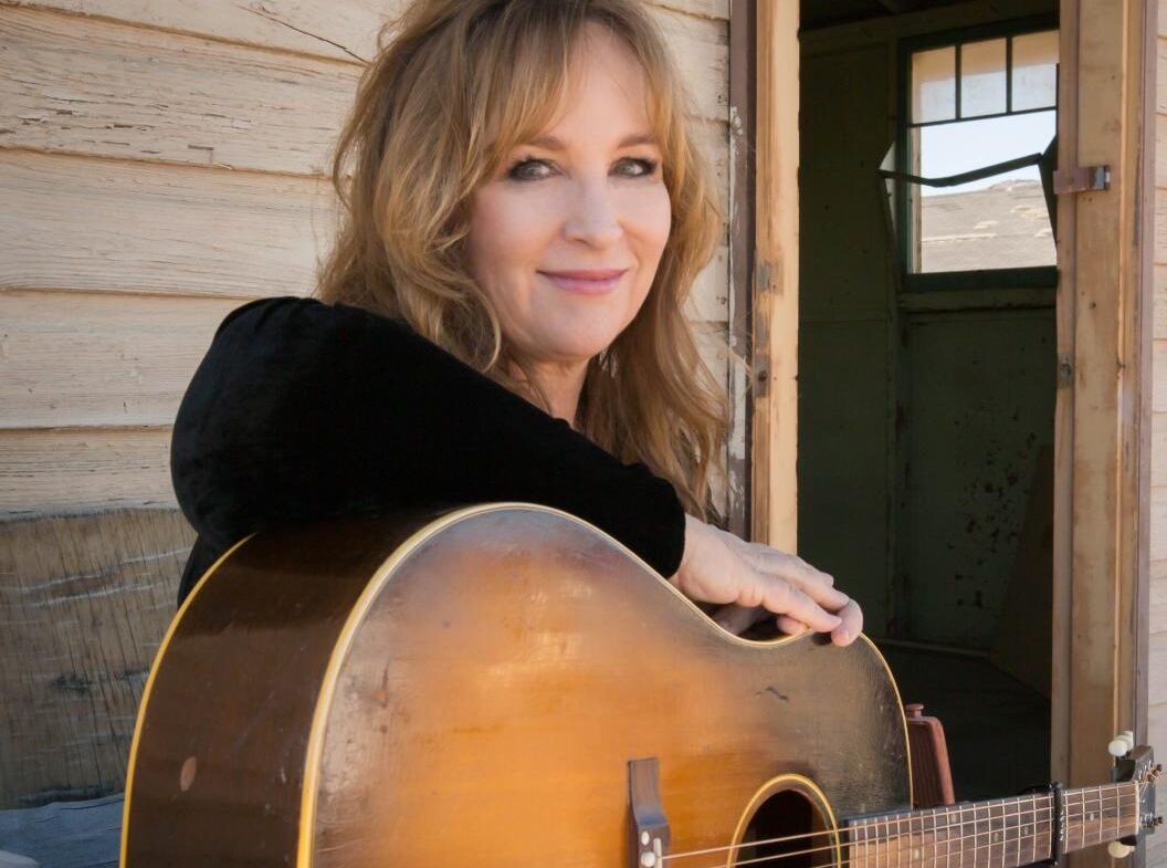 Woman smiling while holding an acoustic guitar near a doorway.