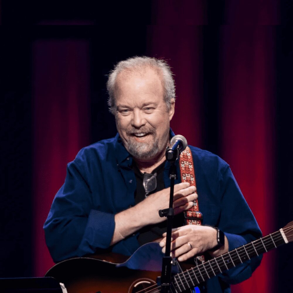Elderly man smiling on stage with guitar and microphone.