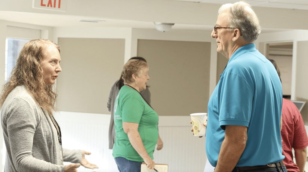 Two elderly people chatting indoors, one holding a cup.