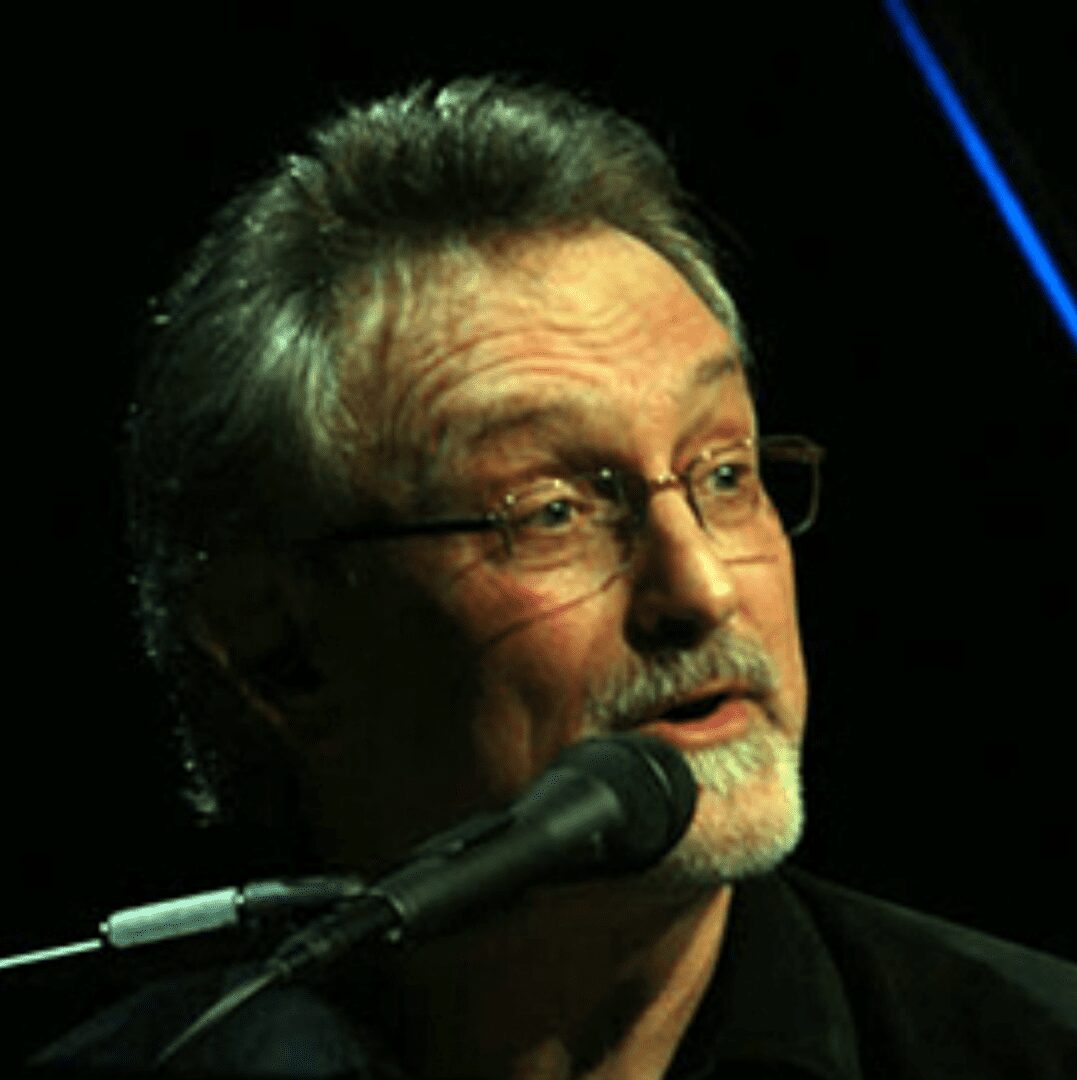 Older man with glasses speaking into a microphone on stage.